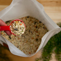 Red measuring scoop filled with mixed grain and herb blend held over open resealable bag of the same mixture