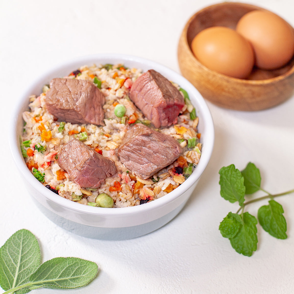 White bowl of beef and vegetable rice with meat chunks, beside brown eggs in wooden bowl and fresh herbs