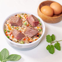 White bowl of beef and vegetable rice with meat chunks, beside brown eggs in wooden bowl and fresh herbs