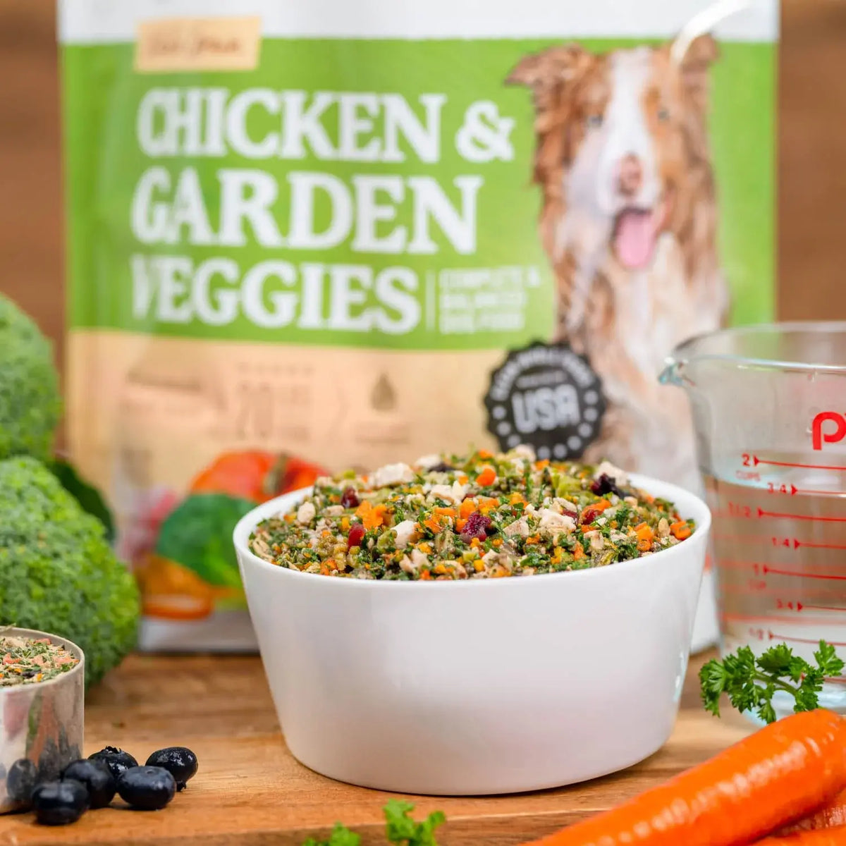 Chicken & Garden Veggies dog food bag beside a white bowl of the fresh food mix, with carrots, broccoli, and blueberries