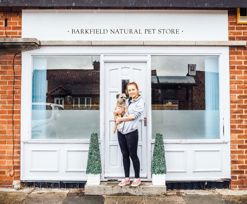 Woman holding a small terrier dog outside Barkfield Natural Pet Store entrance with white shopfront