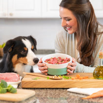 Woman smiling and offering a bowl of raw dog food to a tri-color Australian Shepherd in a kitchen