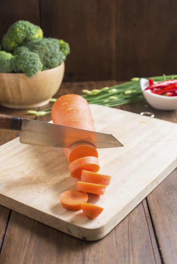 Carrot being sliced on a wooden cutting board with a knife, surrounded by vegetables.