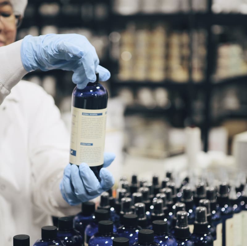 Lab technician in white coat and blue gloves inspecting a blue bottle of herbal shampoo at a production facility