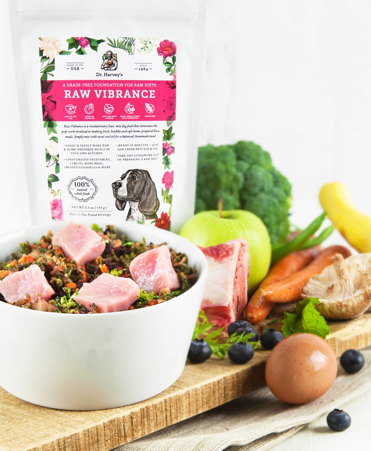 Dr. Harvey's Raw Vibrance grain-free dog food mix bag beside a bowl with raw meat, veggies, egg, blueberries, and bone.