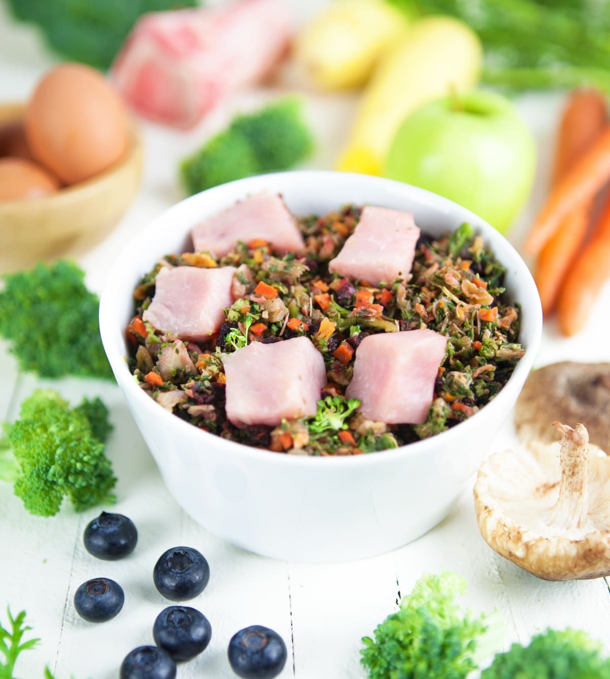 White bowl of fresh dog food with raw meat chunks, mixed vegetables, and greens, surrounded by whole food ingredients.